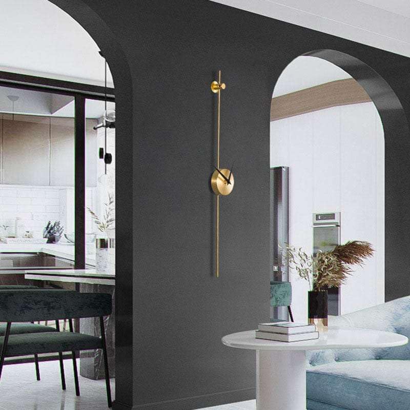 Modern gold wall clock on a dark gray wall with a living room and kitchen in the background.