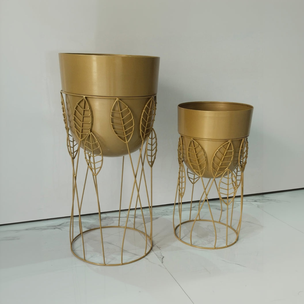 Two gold-colored planters with decorative stands on a white floor.