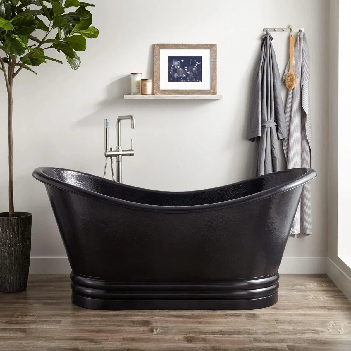 Black freestanding bathtub in a bathroom with a plant, shelf, and towels.