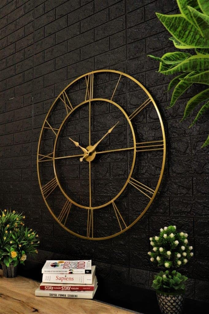 Gold wall clock on a black brick wall with decorative plants and books below.
